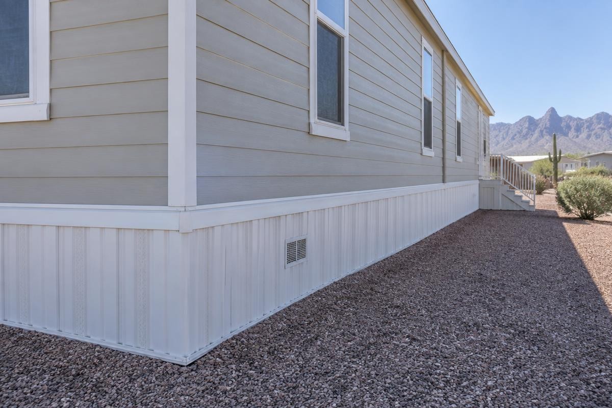 The base of a manufactured home showing a run of white metal skirting panels with a ventilation grille, standing on desert gravel