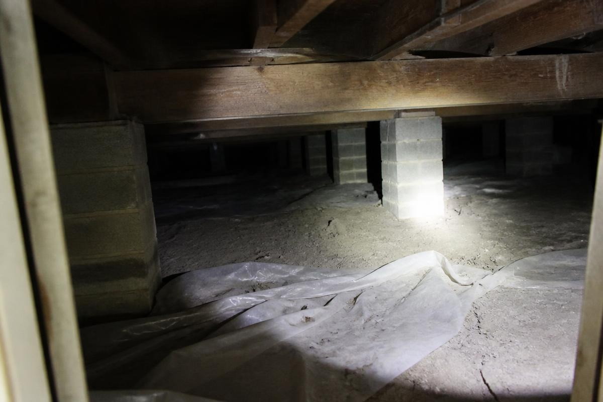 Torchlight in a crawl space showing concrete block piers under timber beams and a torn plastic sheet over bare soil