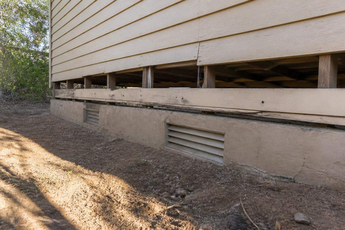 The base of a timber-clad house raised above a concrete foundation wall, with louvred vents and the floor framing visible below the siding