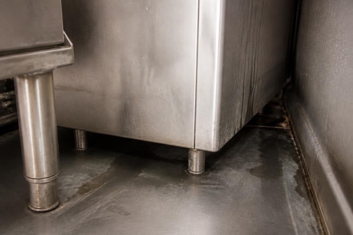 Standing water on the floor beneath stainless kitchen equipment at the wall junction