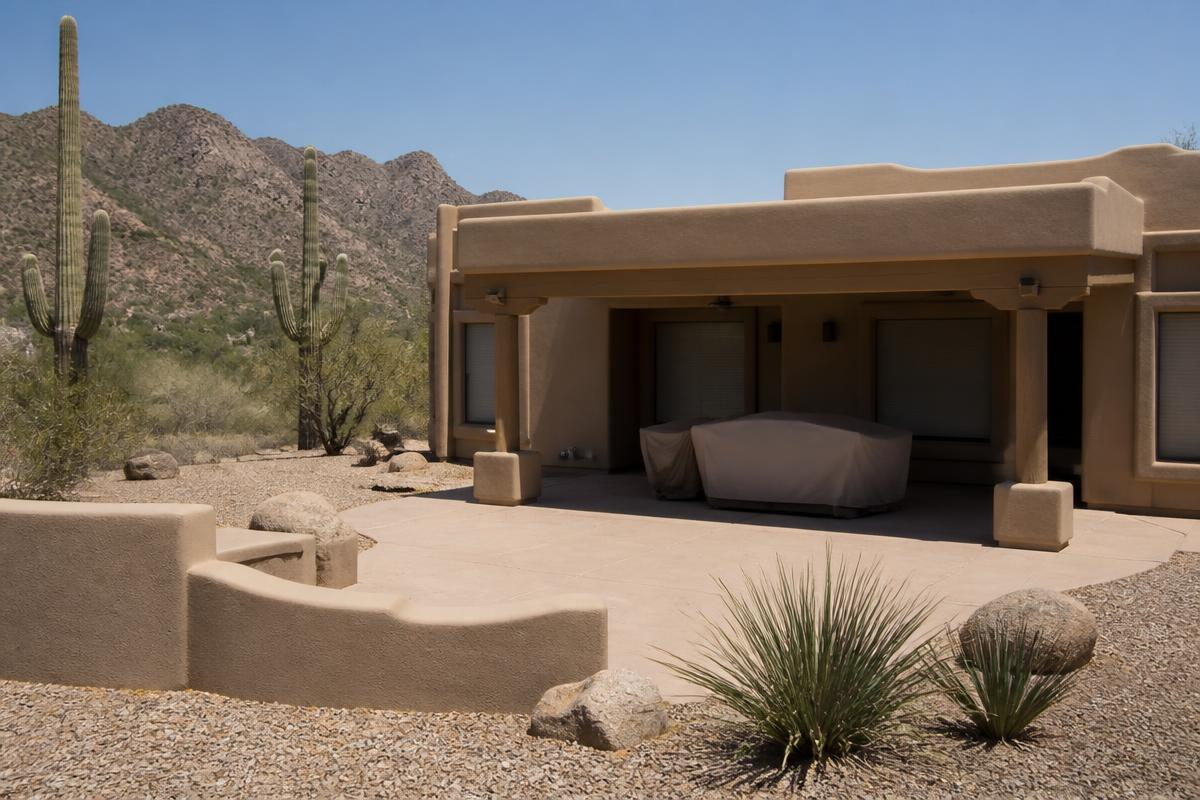 A flat-roofed desert home with blinds drawn and patio furniture under covers, saguaros and a mountain behind