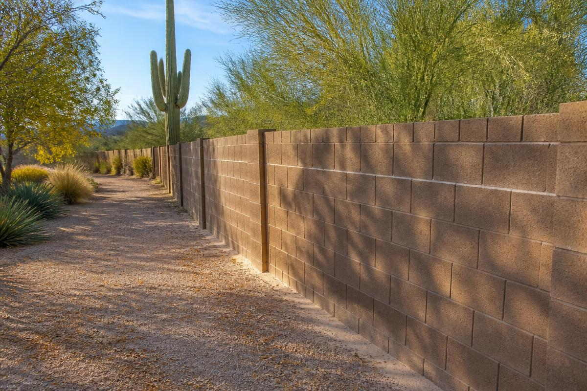 A decomposed granite path running along the base of a long block wall, with a saguaro and palo verde trees behind