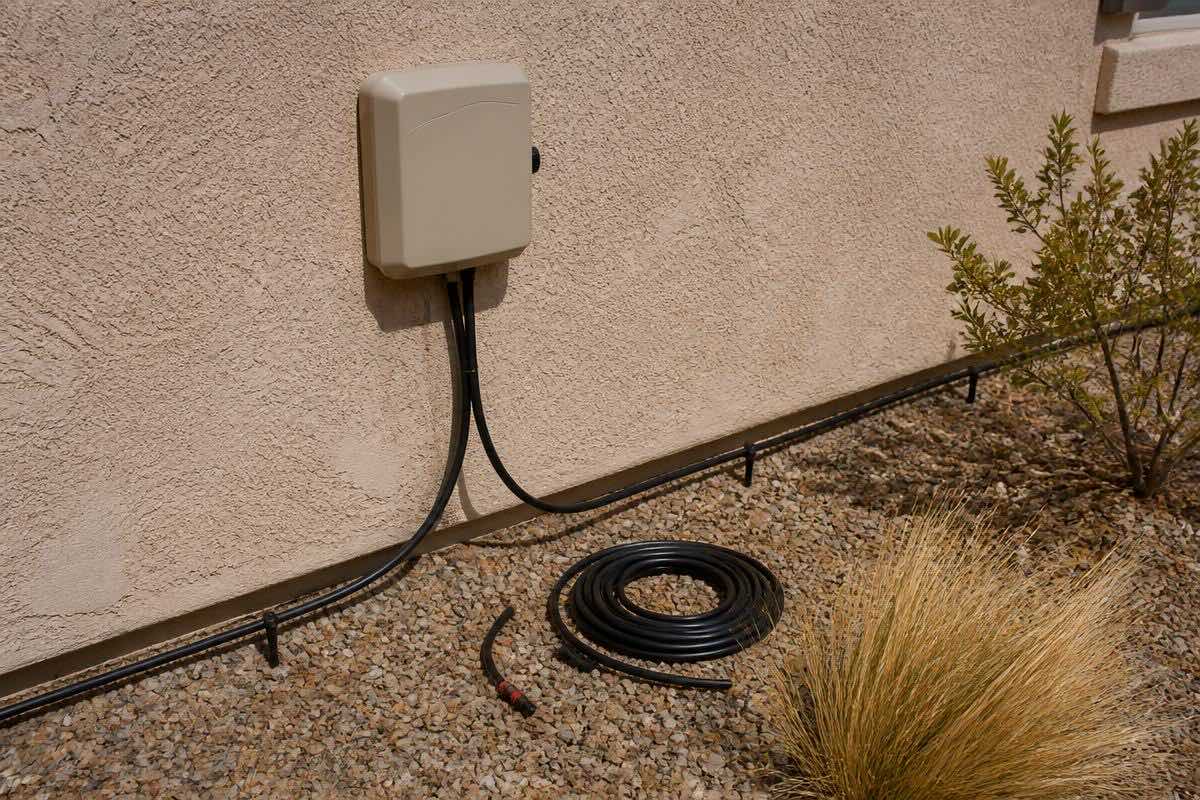A wall-mounted irrigation controller on a stucco wall, with black drip tubing running along the gravel below