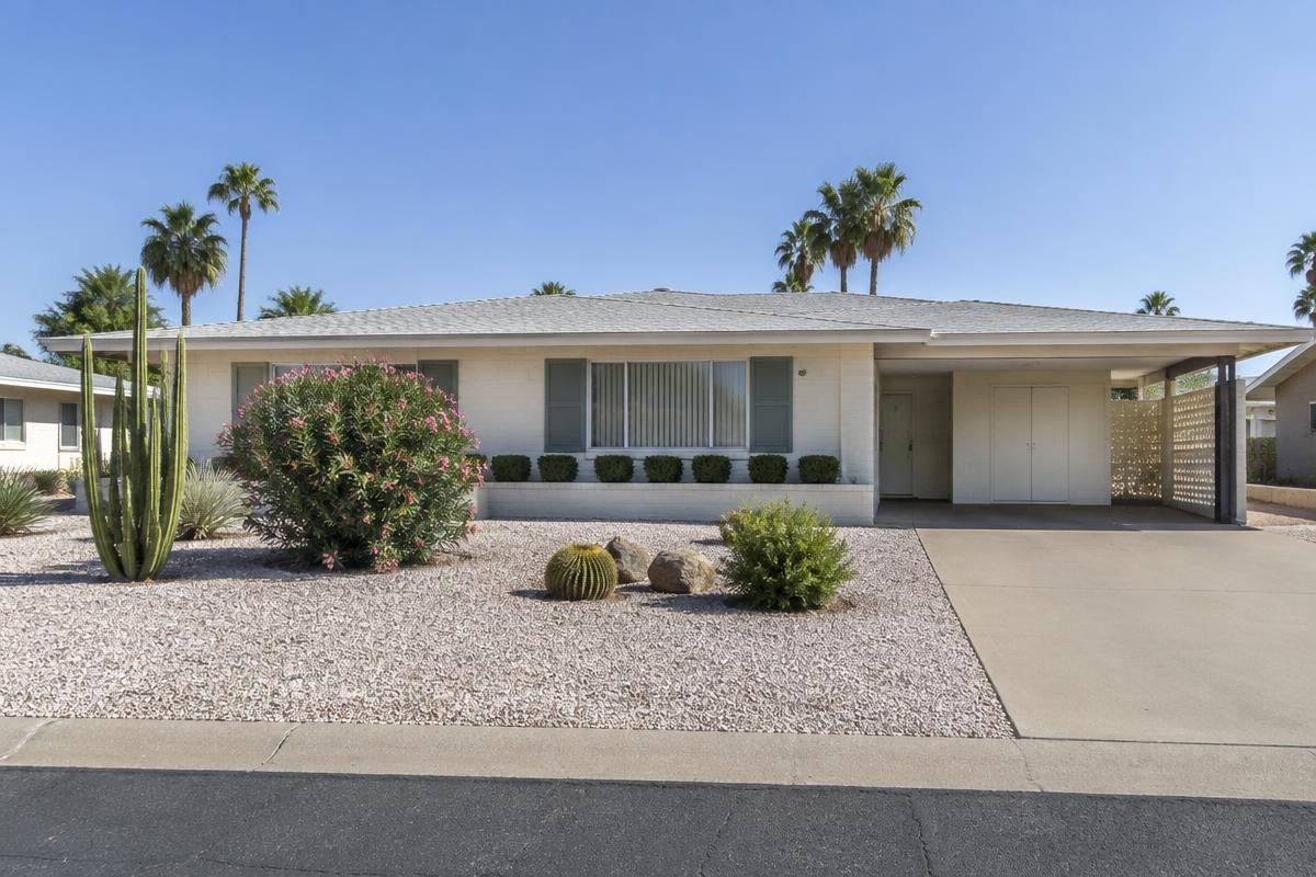 A 1960s single-storey white block house with an open carport, gravel front garden and mature palms behind