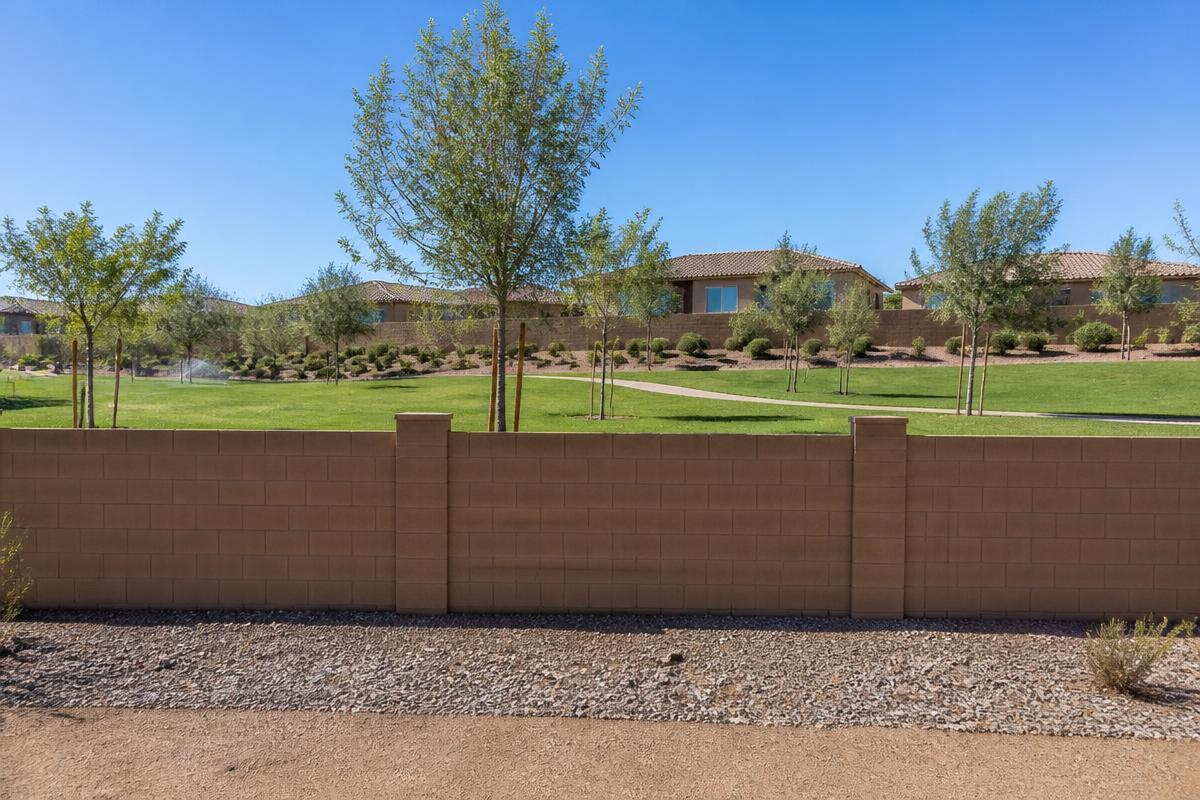 A block wall separating a gravel yard from an irrigated greenbelt with young trees, a path and a running sprinkler, houses beyond