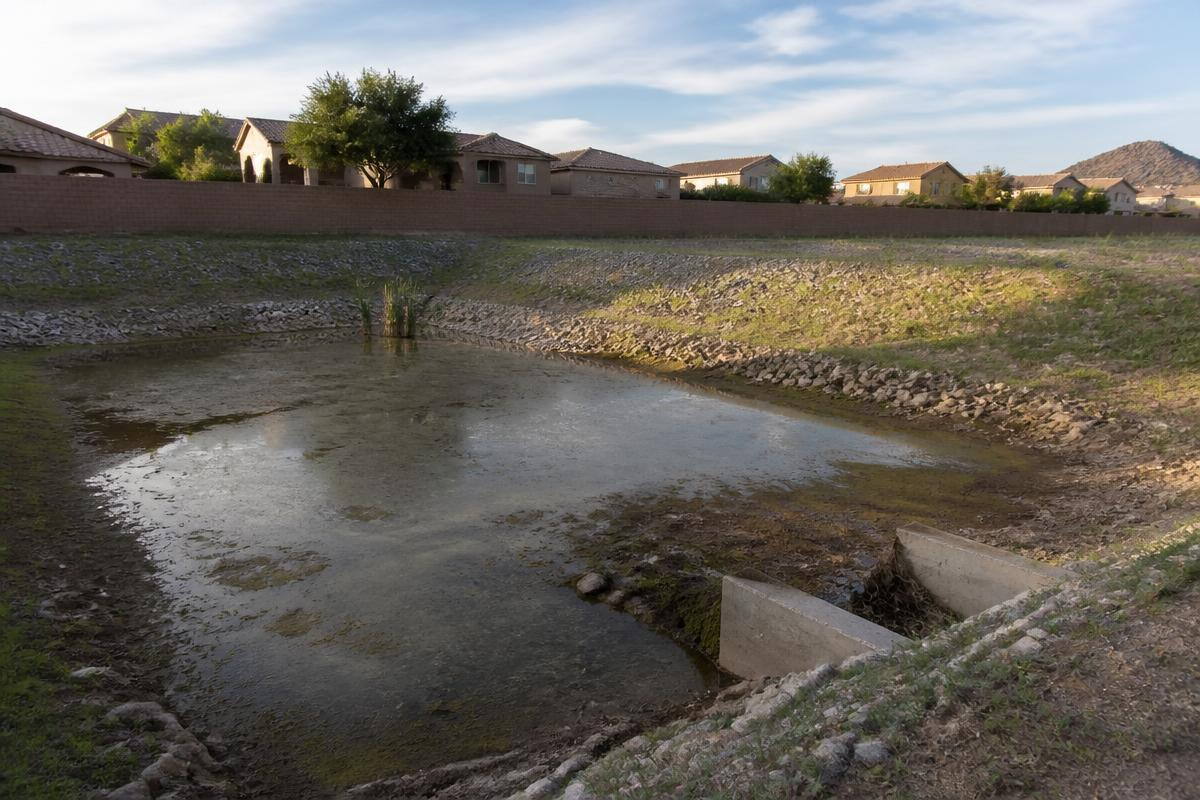 A retention basin holding standing water below a block wall, with a row of tile-roofed houses behind