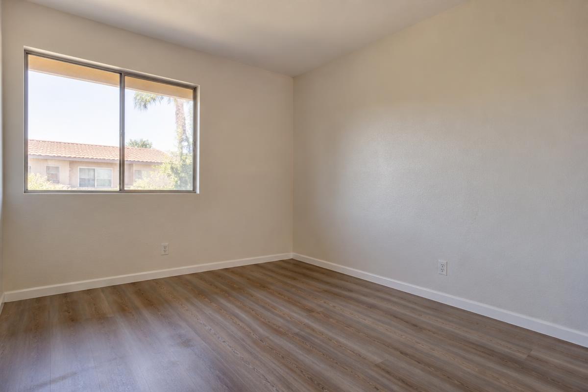 An empty rented room with bare vinyl plank flooring, blank walls and daylight through an uncurtained window looking onto the next building
